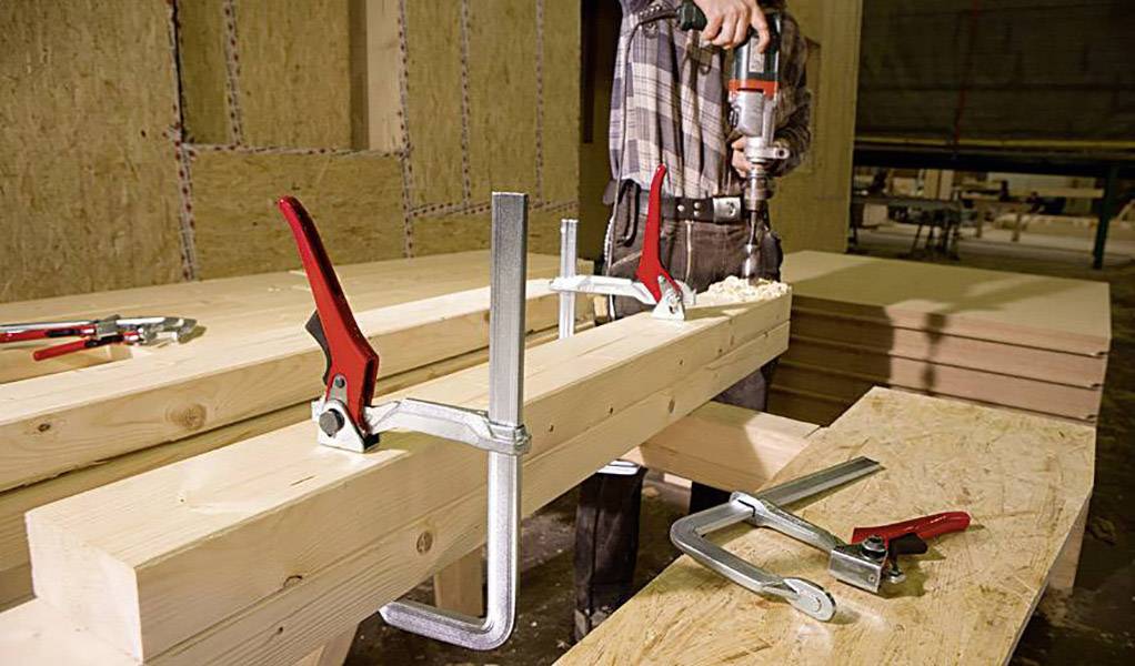 A person is working with wood using a drill in a workshop. Several wooden boards are laid out on two workbenches. G-clamps are present.
