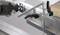 A metal workbench clamp holds a workpiece; a person is operating an electric hand tool for cutting or machining.