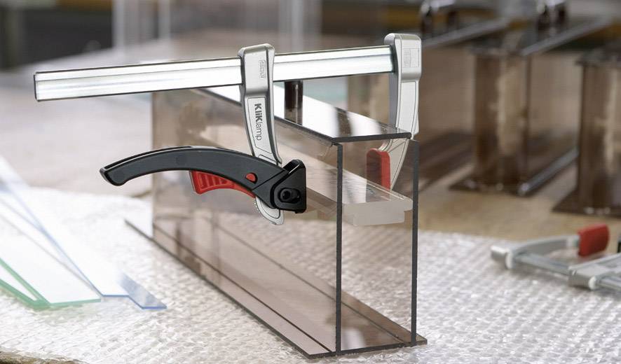 A G-clamp is gripping a transparent plastic part on a padded surface. Tools are scattered in the background.