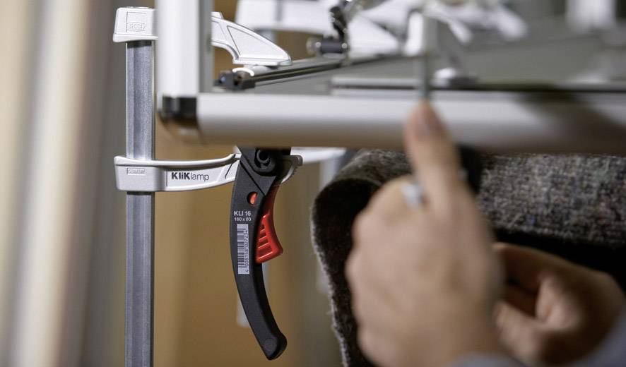 A person is tightening a clamp on a table. The clamp has a black handle with red accents.