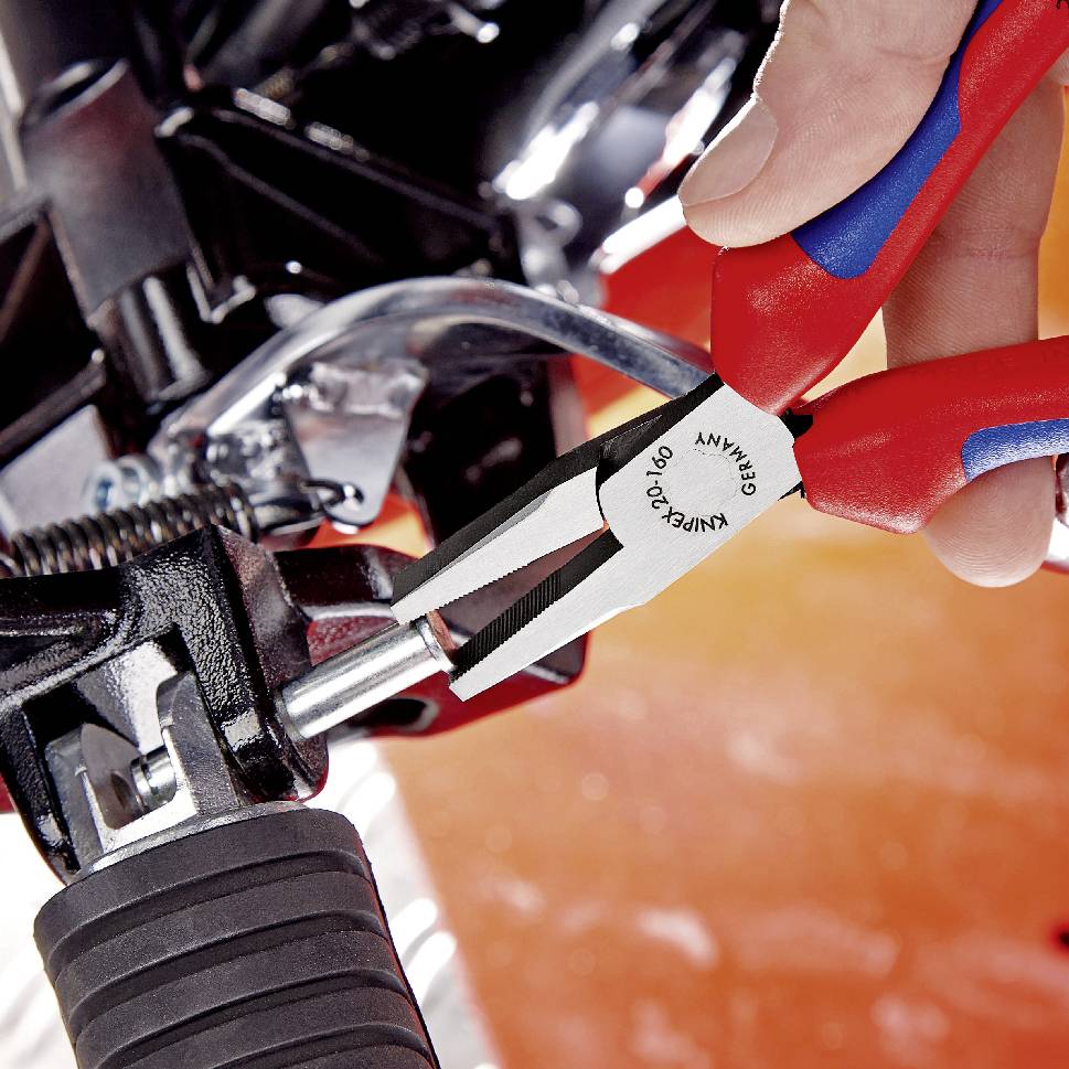 A hand holds a pair of pliers and tightens a screw on a mechanical device. The pliers are red and blue and made in Germany.