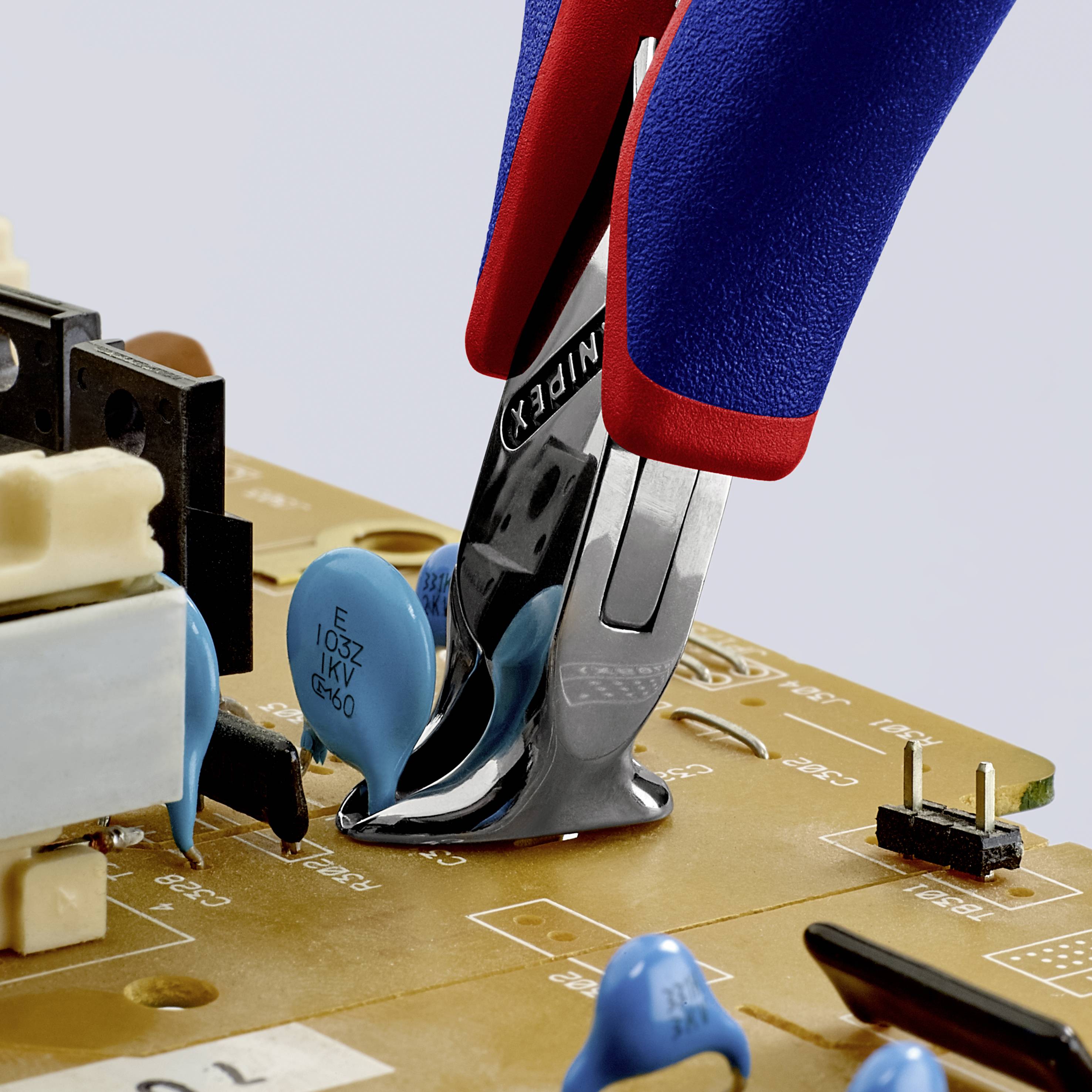 A hand holds a red and blue pair of pliers, cutting a wire on a circuit board with various electronic components.