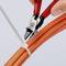 A hand is cutting a white cable tie with a red pair of pliers, which is wrapped around several orange cables.