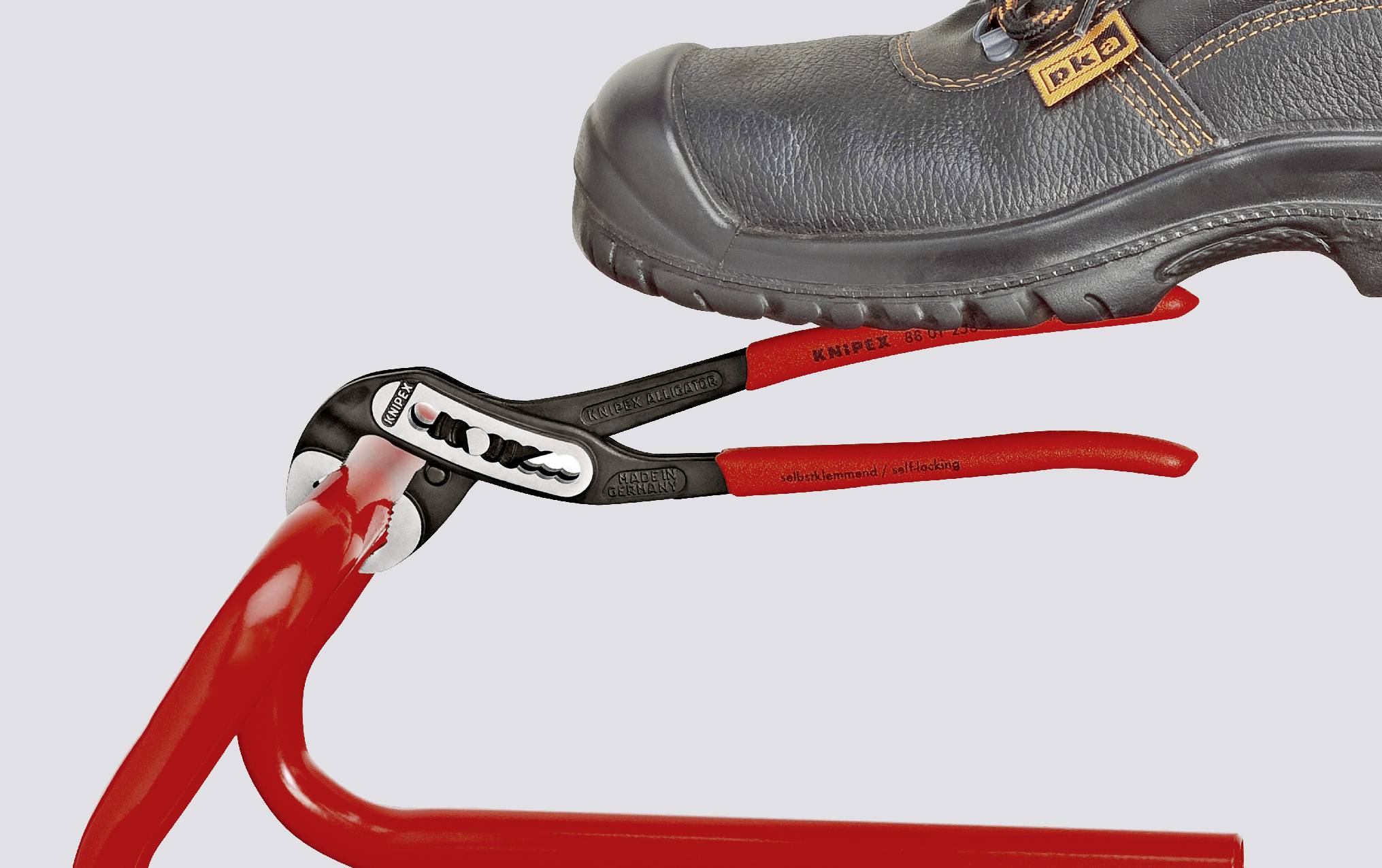 A black safety shoe is holding a red pipe with a red pair of pliers.
