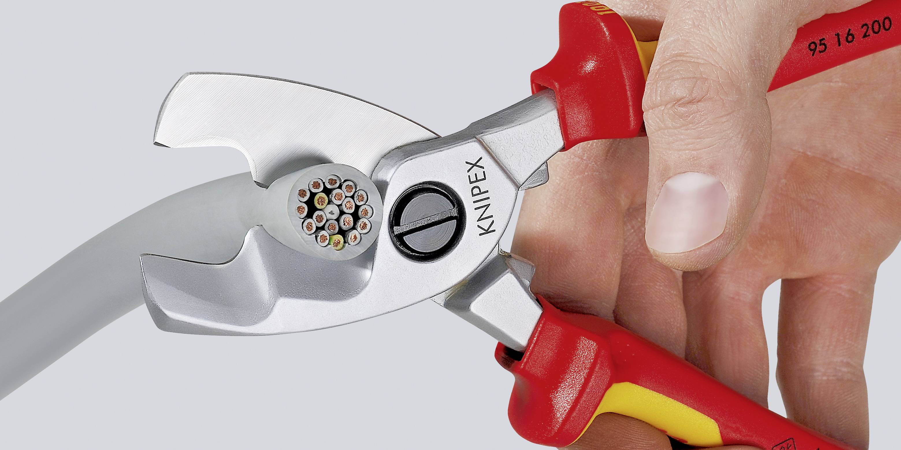 A hand is holding a red and yellow cable cutter pliers, cutting through a cable with multiple wire cores.