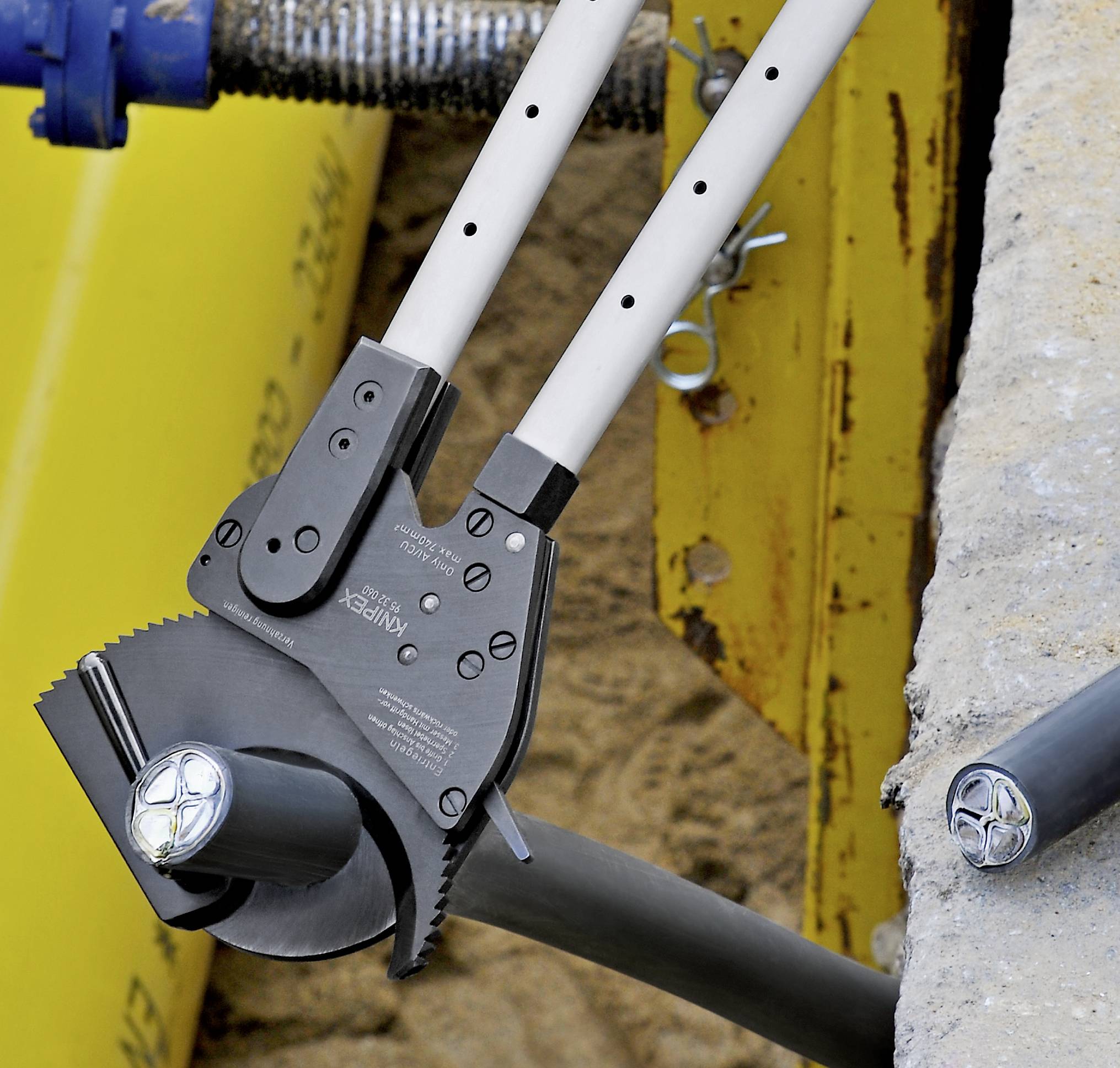 A wire cutter is cutting a thick cable. Yellow and metallic construction elements are visible in the background.