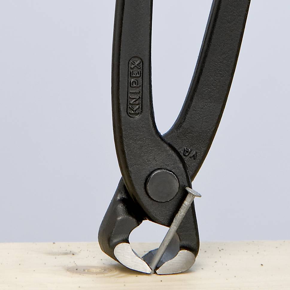 A black pair of pliers cuts a wire on a wooden block. The pliers are positioned vertically and point downwards.