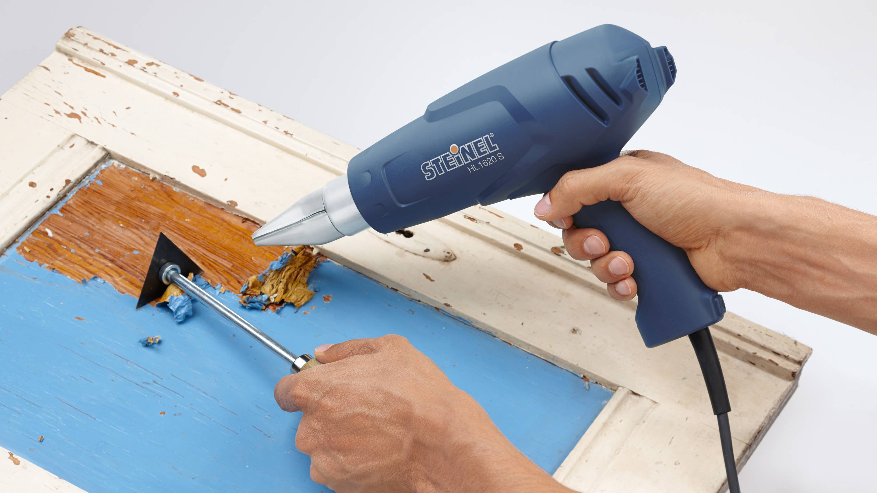 Two hands are operating a hot air blower and a scraper to remove blue paint from old wood.