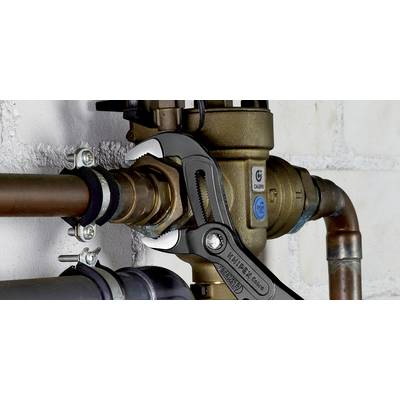 An adjustable pipe wrench firmly grips a metal joint in a piping system mounted on a white brick wall.