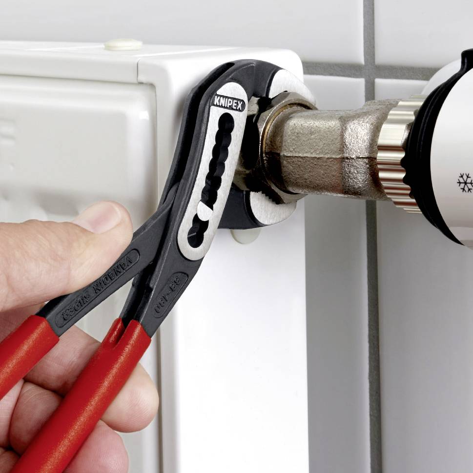 A hand tightens a heating valve connection with a red pair of pliers to secure the seal. Wall tiles in the background.