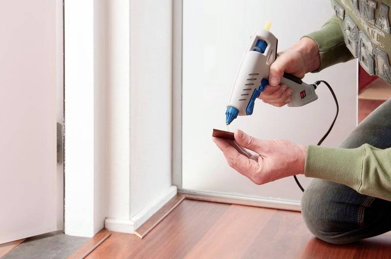 A person is gluing a door threshold with a hot glue gun. The floor is made of wood laminate and the person is wearing a green top.