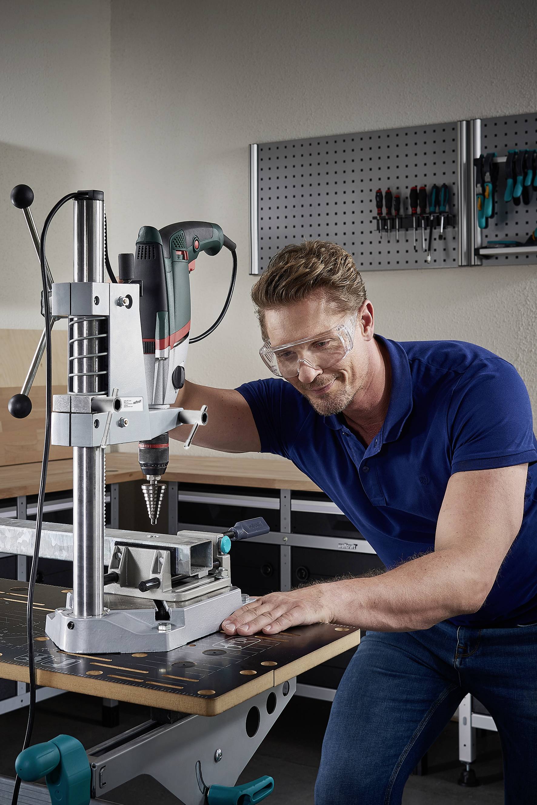 A man is using a bench drill in a workshop. He is wearing safety glasses. Tools are hanging on the wall in the background.