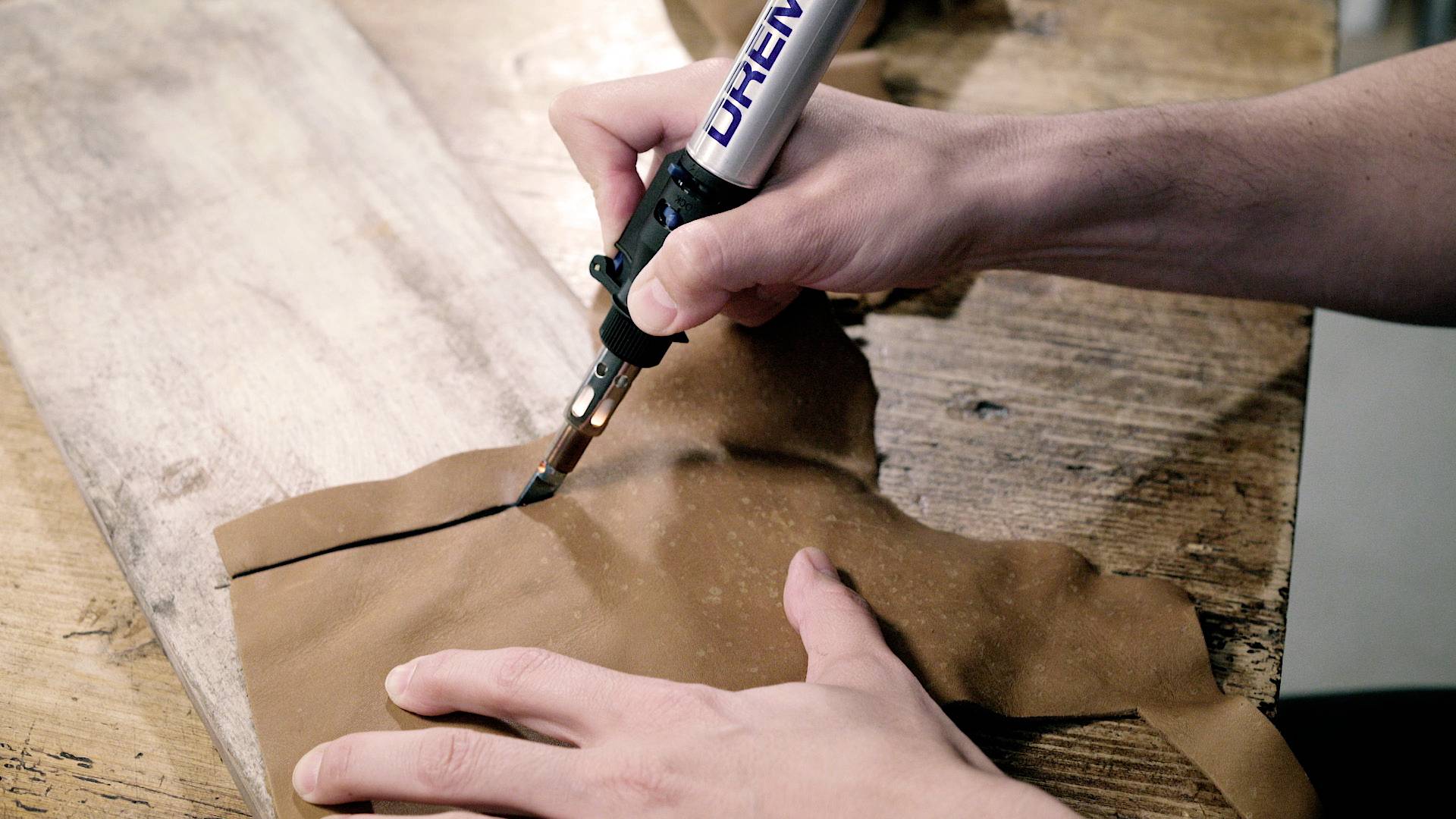 A person is cutting leather with a hot tool on a wooden table.