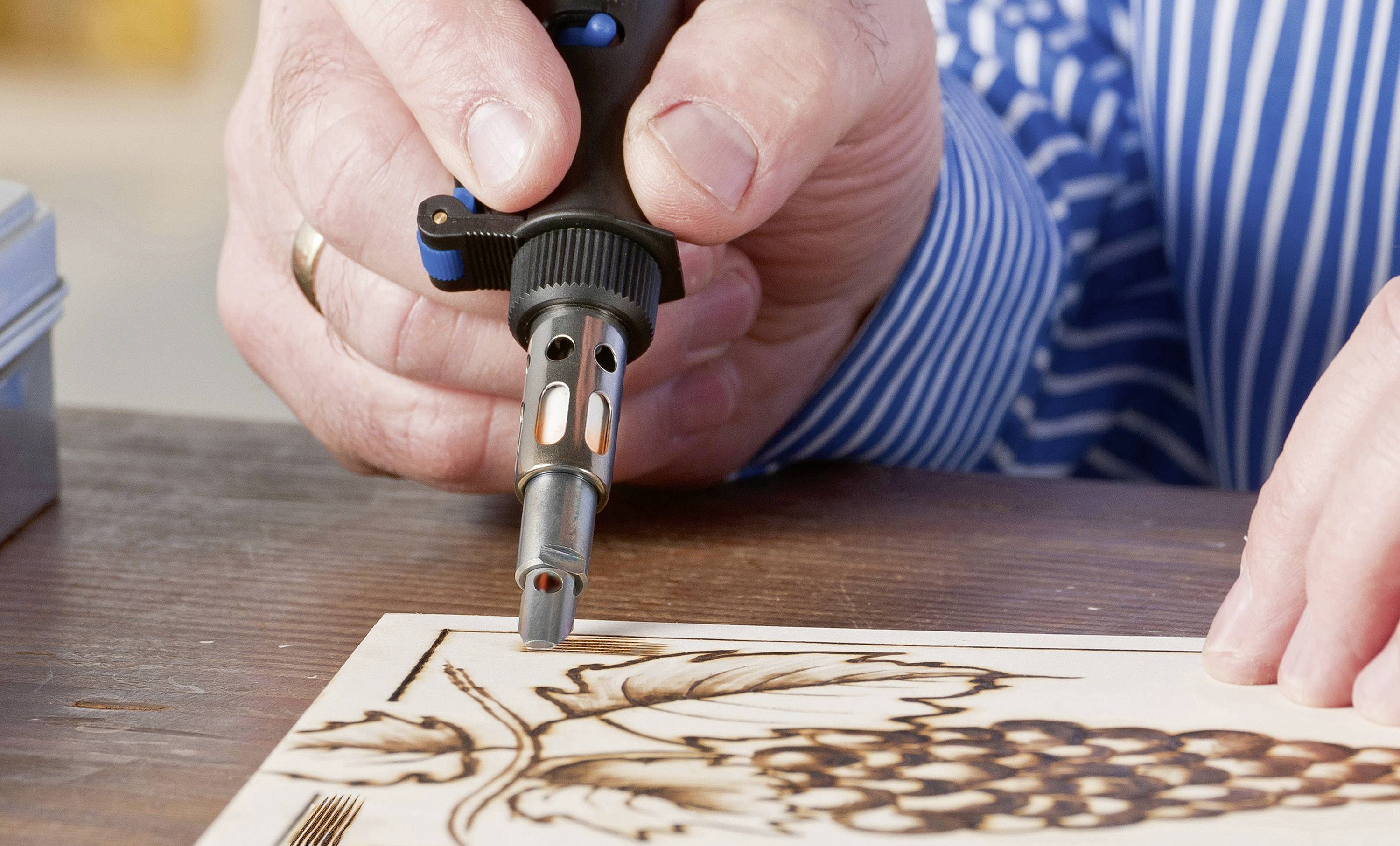 A person is using a burning tool to engrave grape patterns onto wood. The person is wearing a blue and white striped shirt.