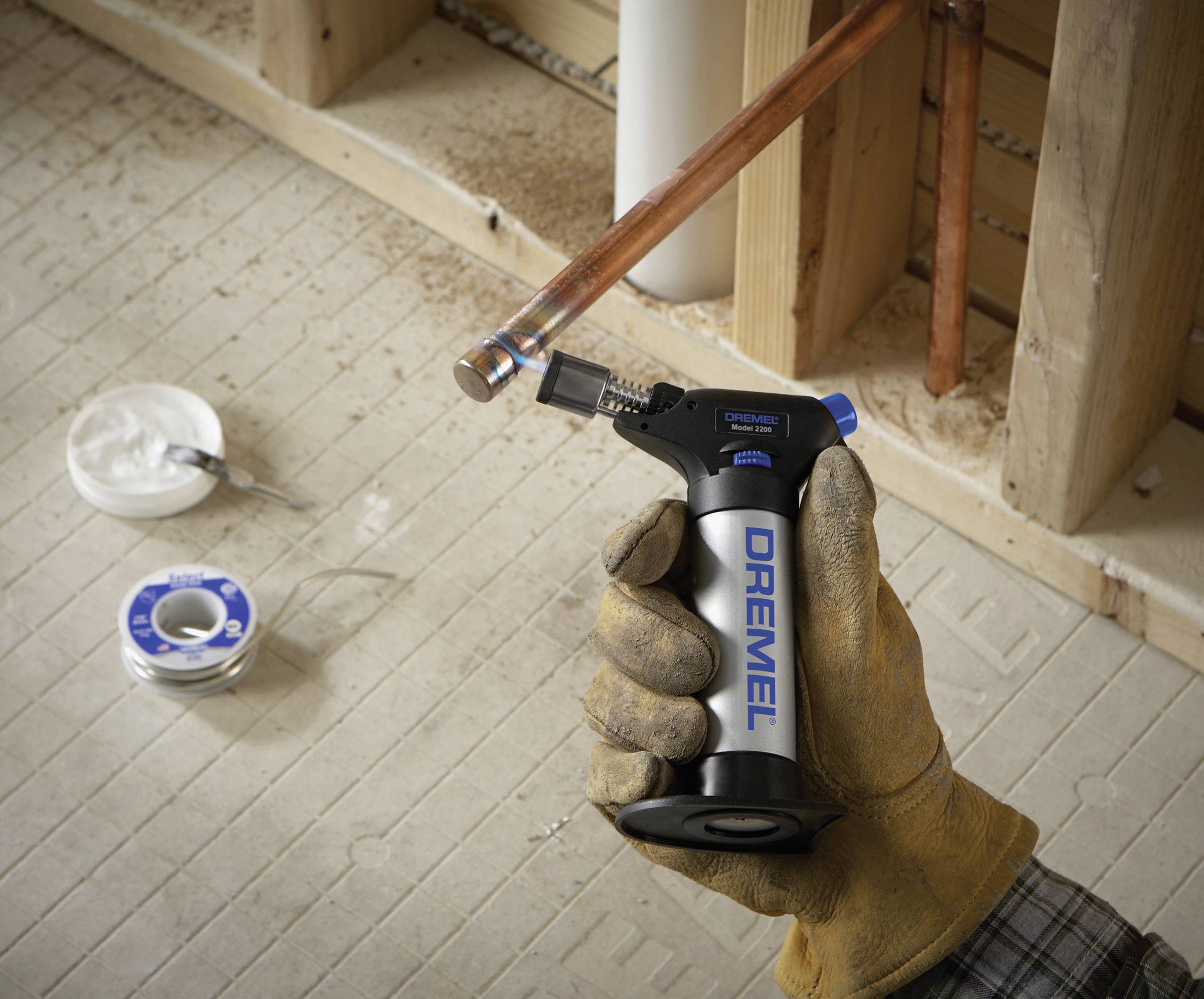 A person wearing gloves holds a small hand torch in front of a copper pipe, ready to solder. Soldering materials are visible in the background.