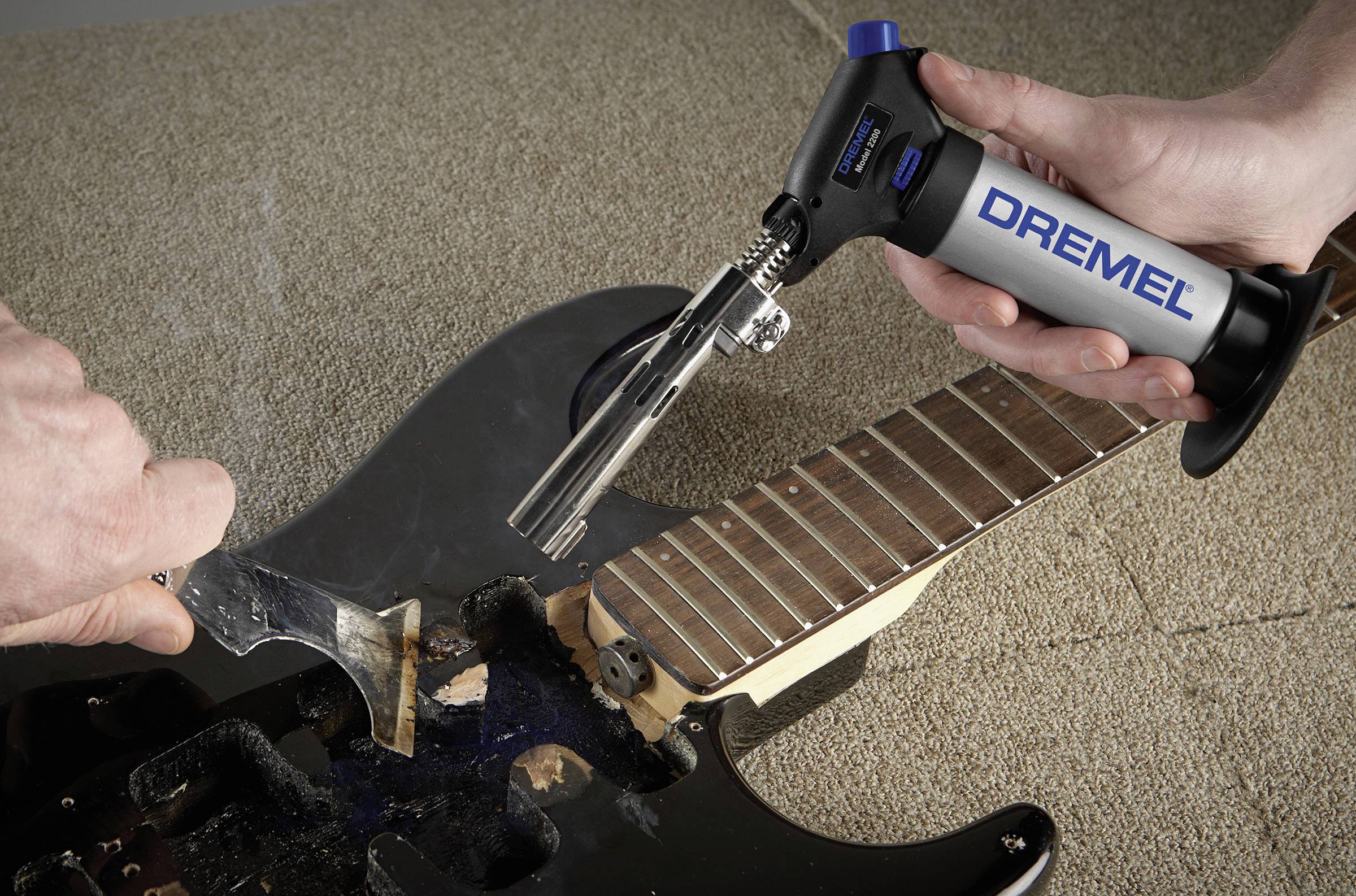 A person is working on a black electric guitar with a Dremel tool. The neck of the guitar remains untouched while the body is being processed.