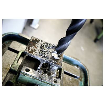 Close-up of a drill bit boring into a metal block clamped in a vise, with metal shavings around the hole.