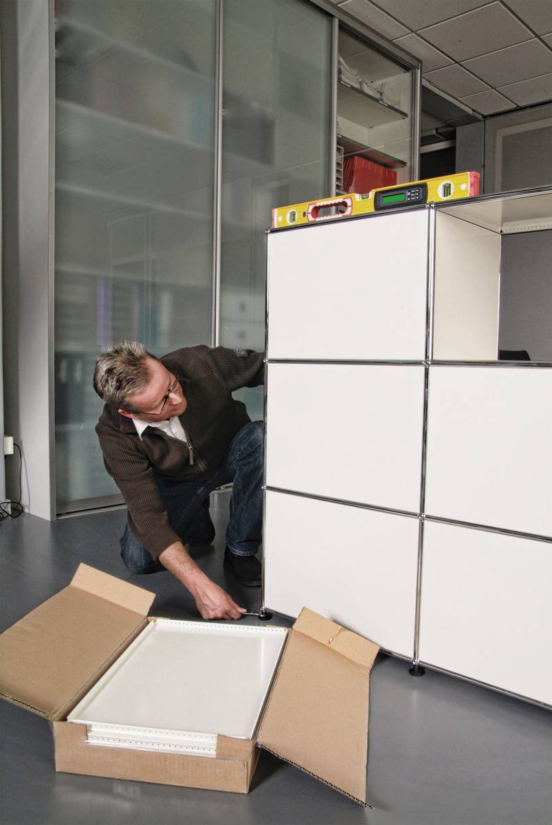 A man is positioning a cabinet using a spirit level and a water bottle. An open cardboard box is lying in front of it.