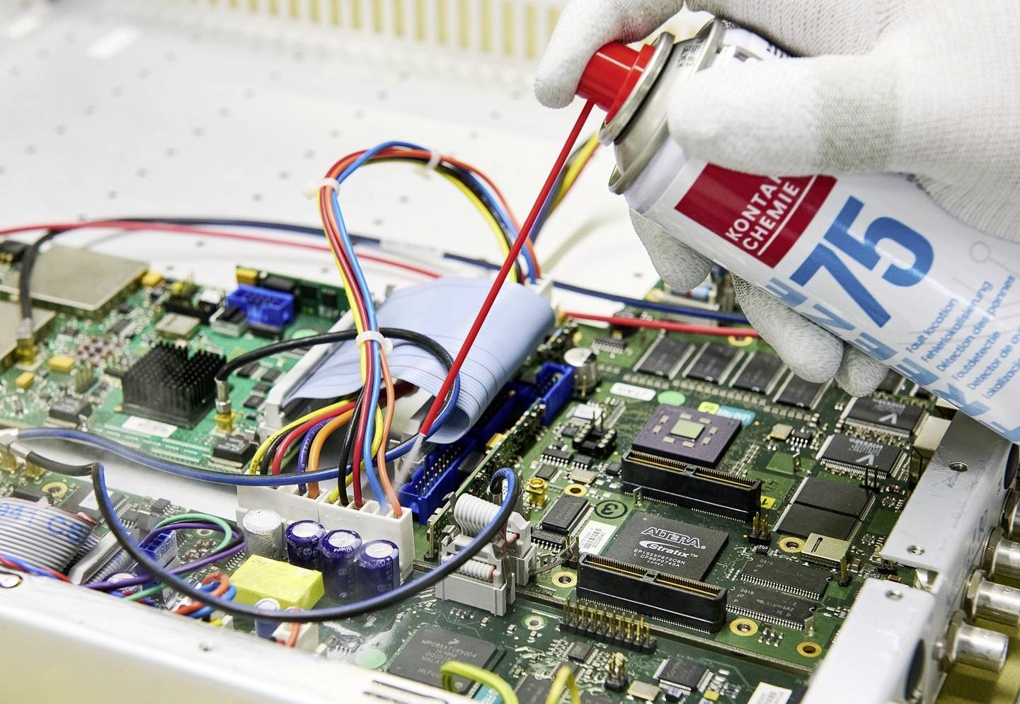 A gloved hand sprays coolant from a can onto the circuits of an open computer circuit board with colourful cables.