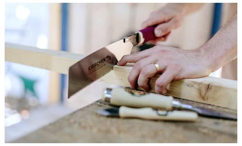 Close-up of hands using a hand saw to cut a wooden board on a workbench, with tools in the foreground.<br>