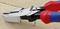 Pliers with red and blue handles are cutting a nail into a piece of wood. The metal jaws have teeth for a firm grip.