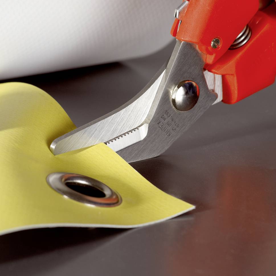 A red pair of scissors is cutting a yellow material with a reinforced eyelet, possibly for fastening, against a neutral background.