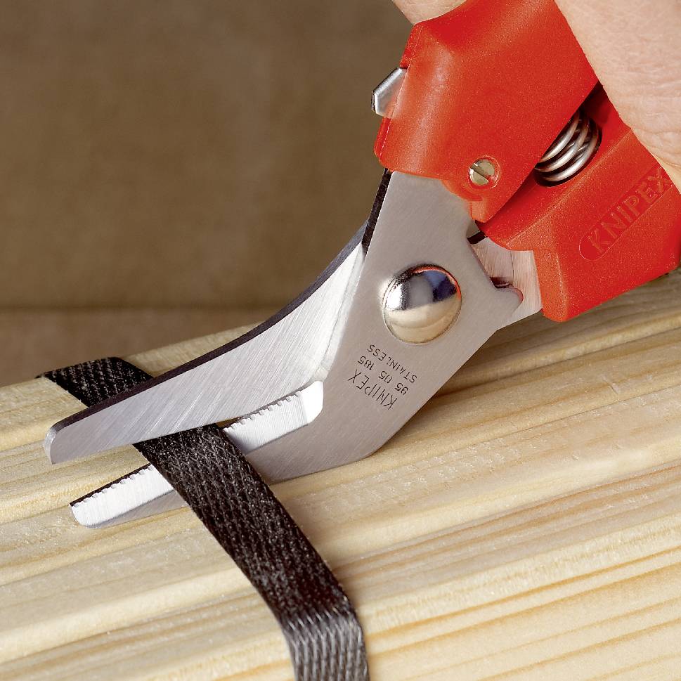 A red scissors cuts through a black elastic band on a wooden board.