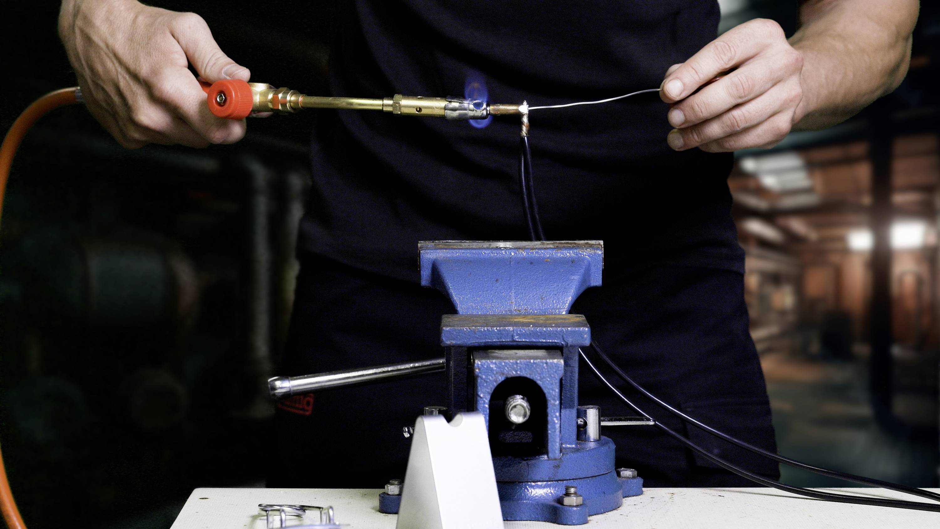 A person is soldering a piece of wire with a soldering iron at a vice in a workshop environment.