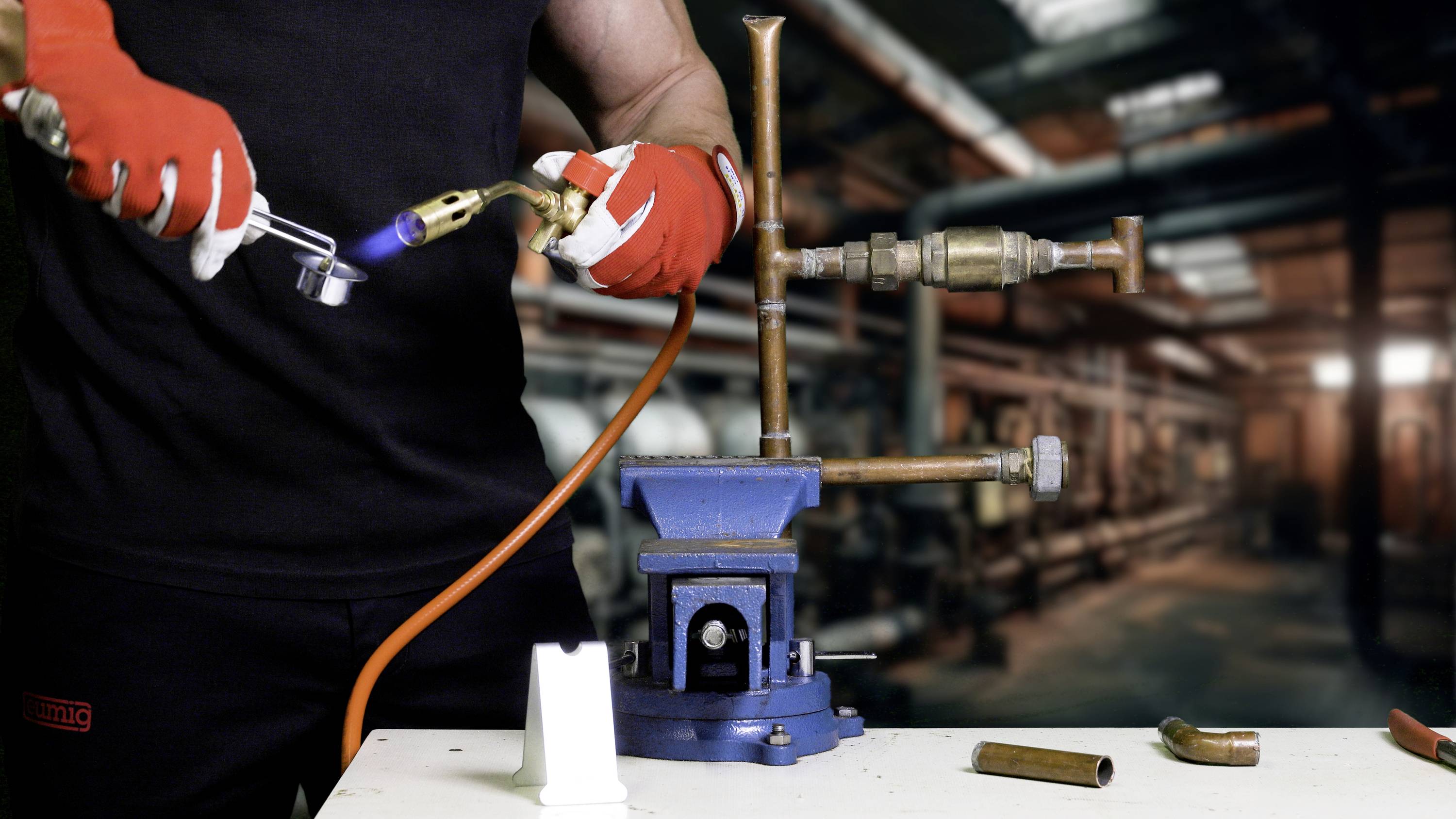 A craftsman in black clothing is using a welding torch to work on a copper pipe in a workshop environment.