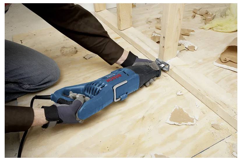 A person uses an electric oscillating tool to trim a wooden board in a construction setting. Pieces of debris are scattered around.