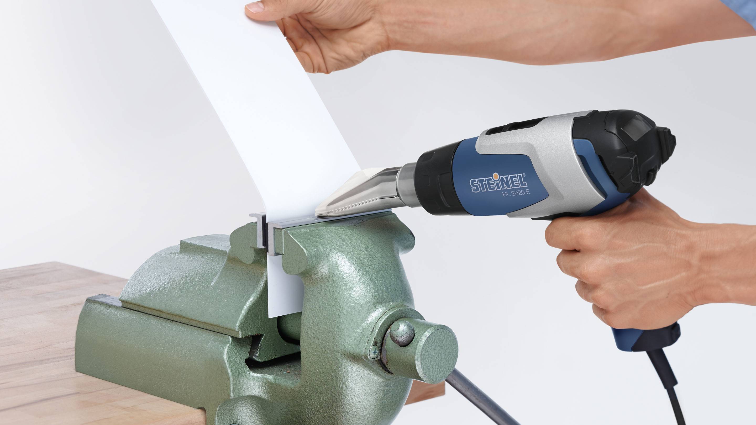 A person is holding a heat gun and working on a white sheet metal that is clamped in a vice.
