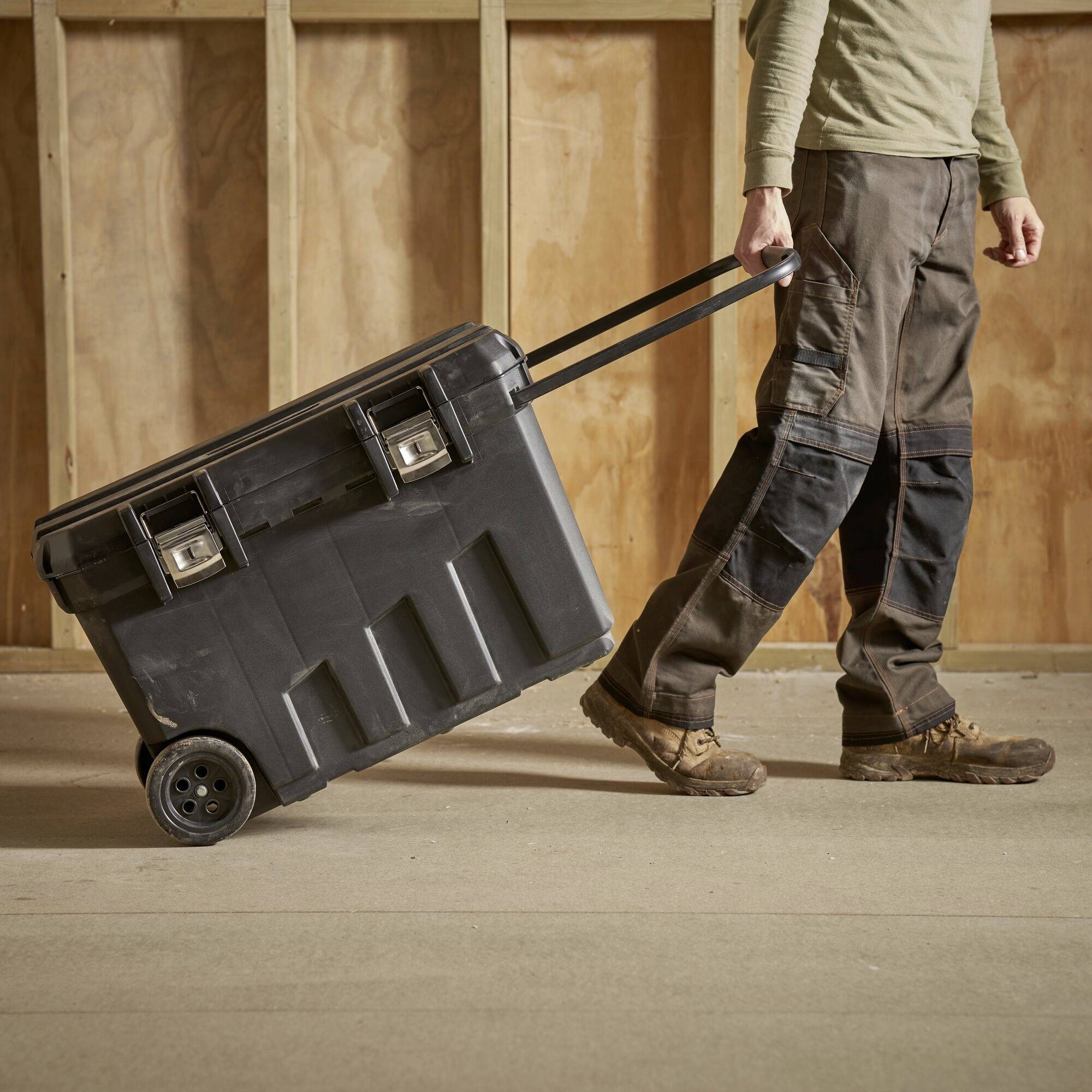 A person is pulling a large, black toolbox with wheels along a wooden frame. They are wearing a work uniform.