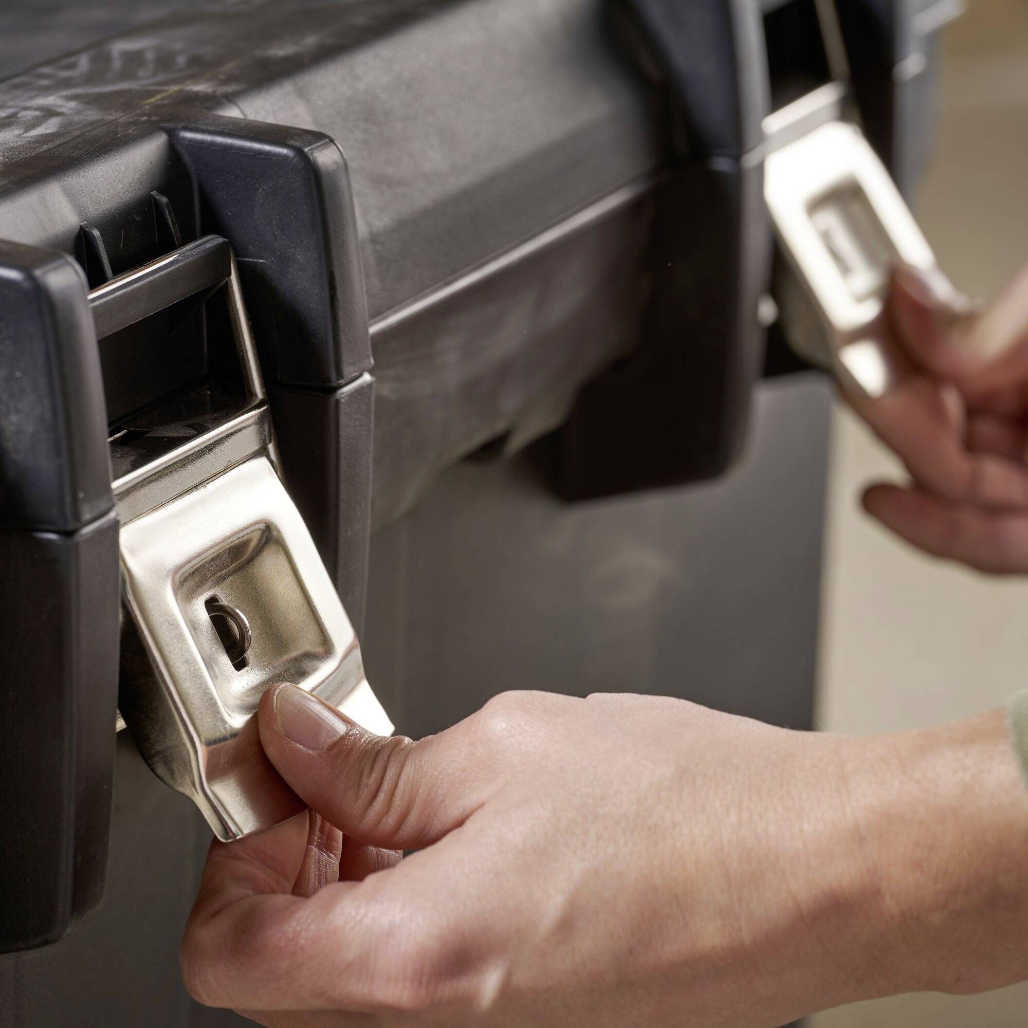 A hand reaches for a metal fastener attached to a robust container, possibly a waste bin.