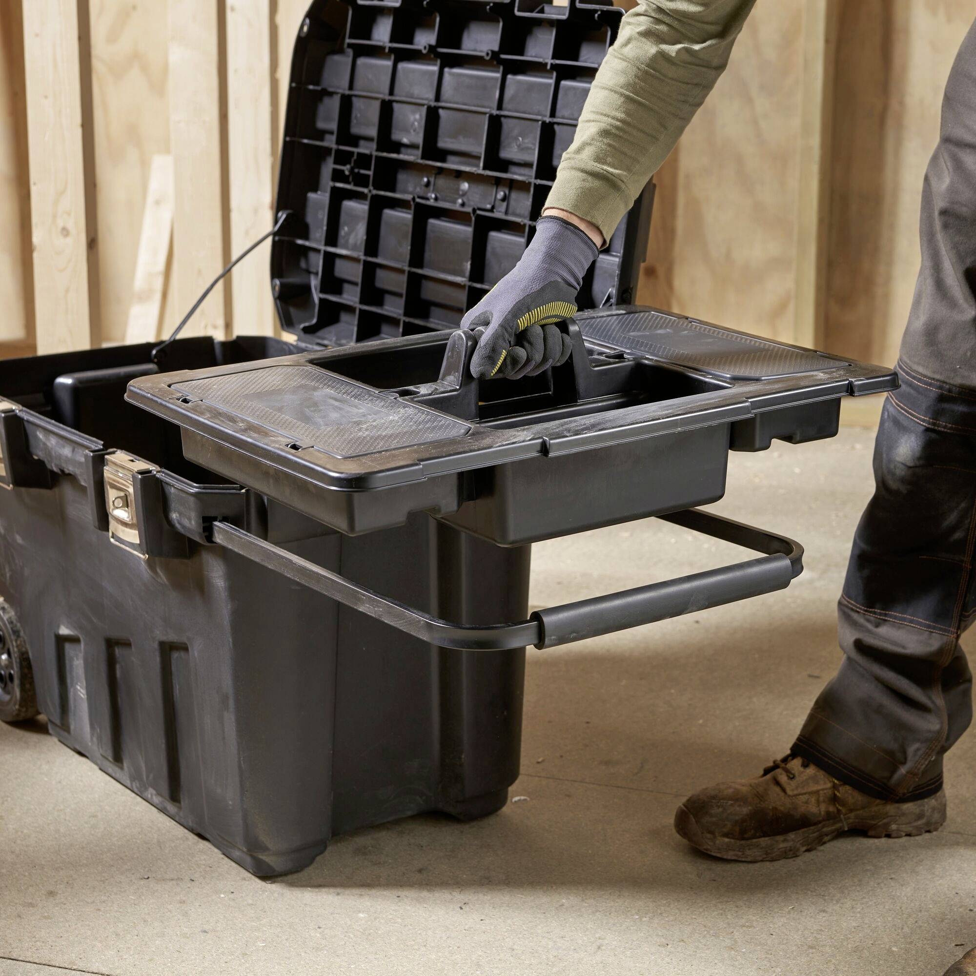 A person is using a toolbox with multiple compartments in a workshop. The person is wearing work gloves and work boots.