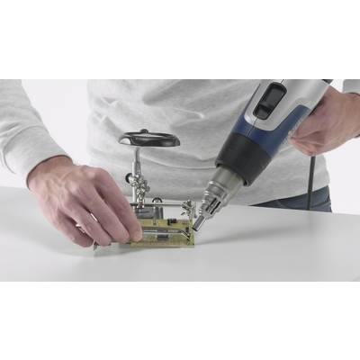 A person is soldering a circuit board with a hot air gun. They are holding the board on a stand while the device is in their other hand.