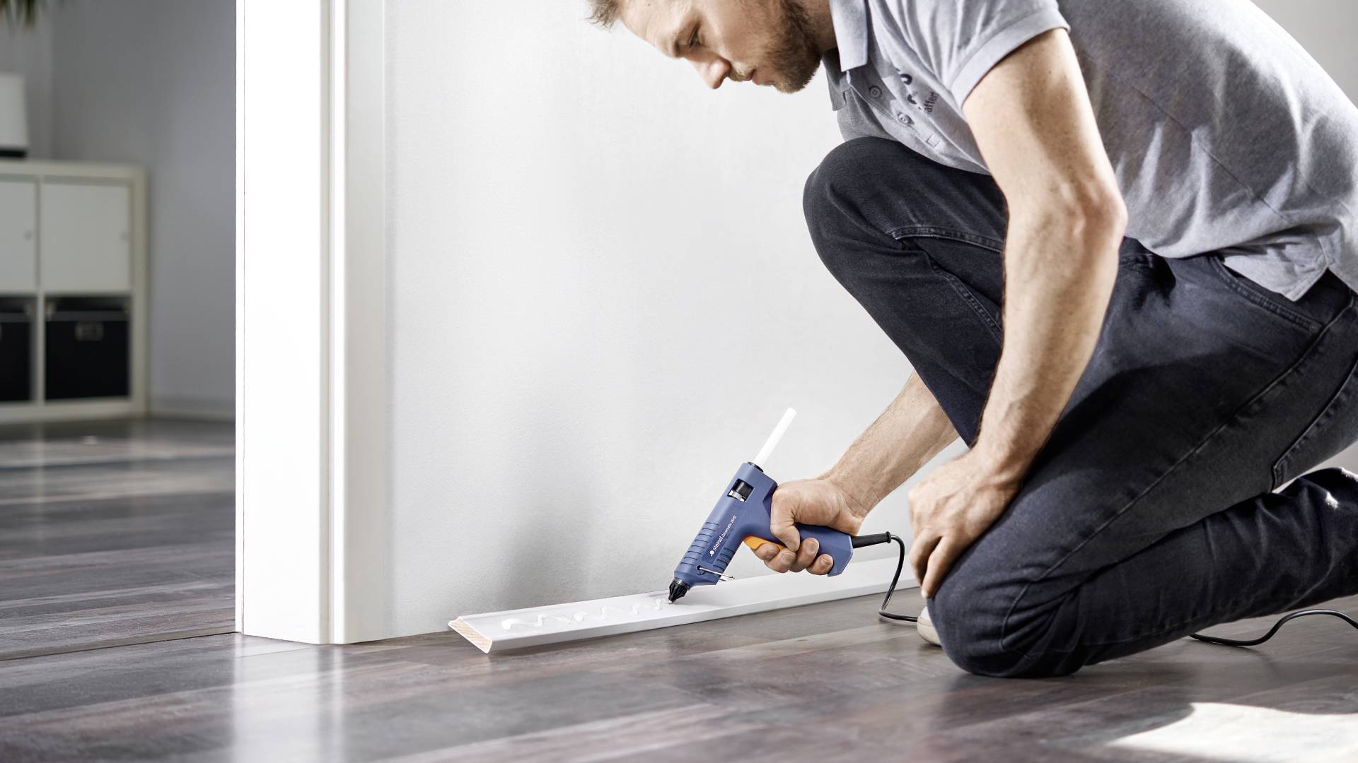 A man is fixing a white skirting board to the floor with a hot glue gun. He is kneeling on a laminate floor in a modern room.