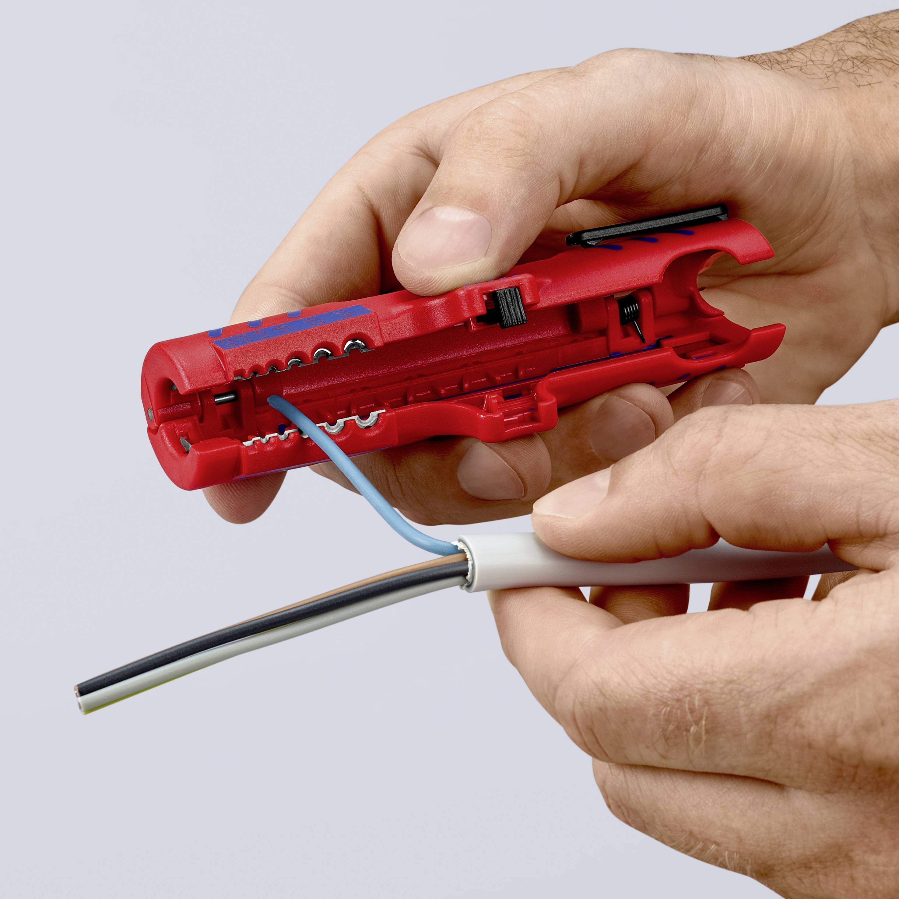 A hand is using a red wire stripping tool to remove the sheathing of a cable.