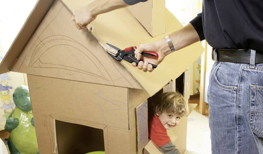 A person is building a cardboard house, while a child looks out cheerfully from a window.