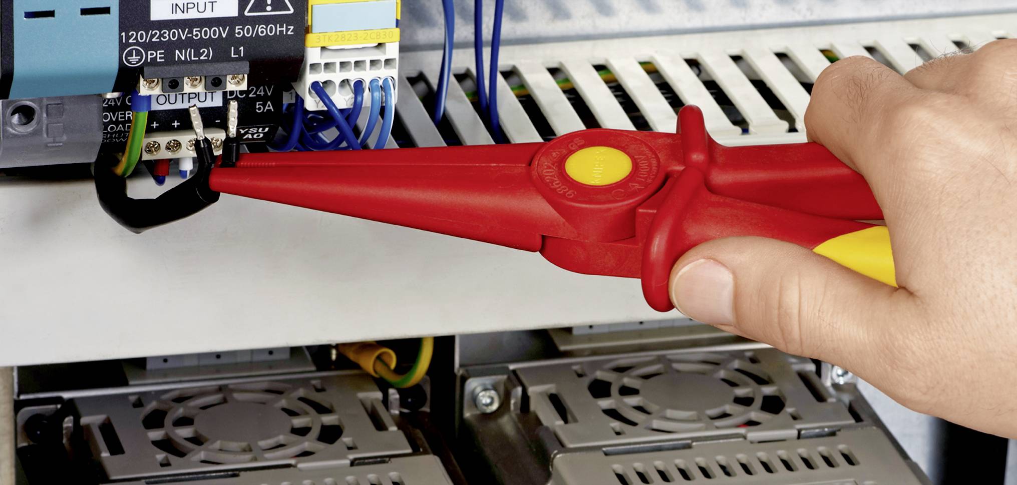 A red plier holds the black cable on an electrical device in a control cabinet. Safety inspection is being carried out.