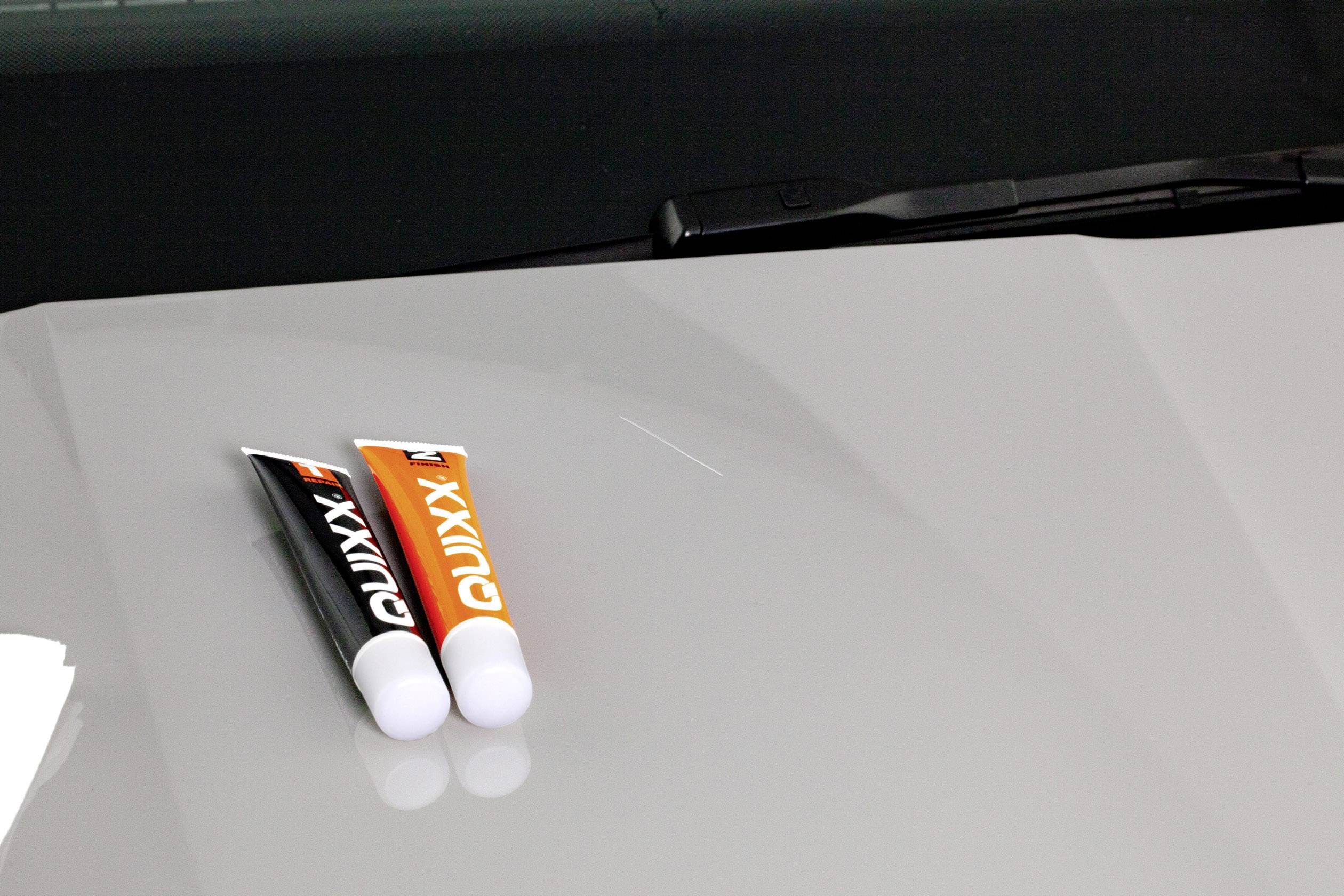 Two tubes of polish with the label 'Quixx' are lying side by side on a grey car body. A windscreen wiper is visible in the background.