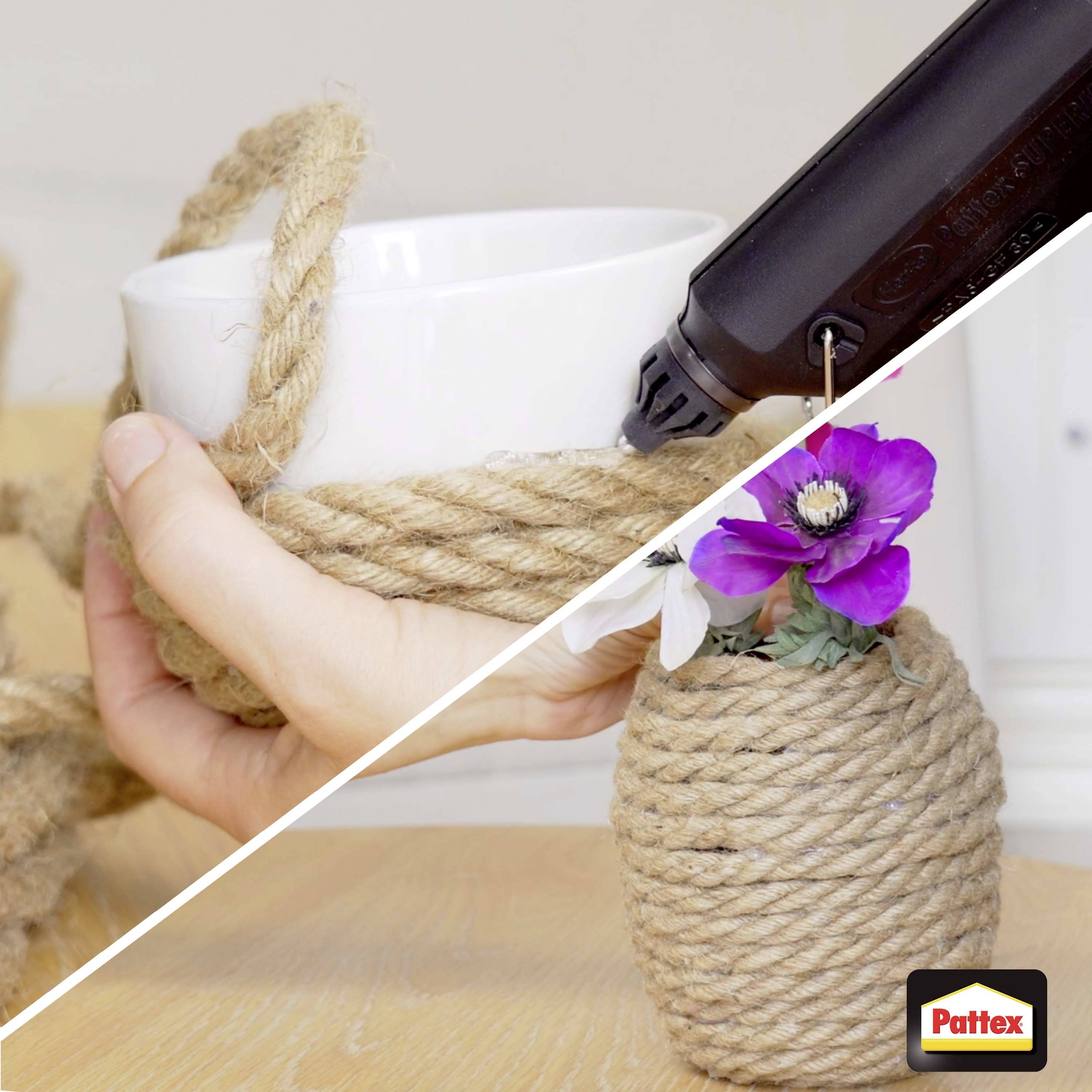 'Hand wrapping a rope around a white cup with a hot glue gun; cup decorated with a purple flower, set on a wooden table.'