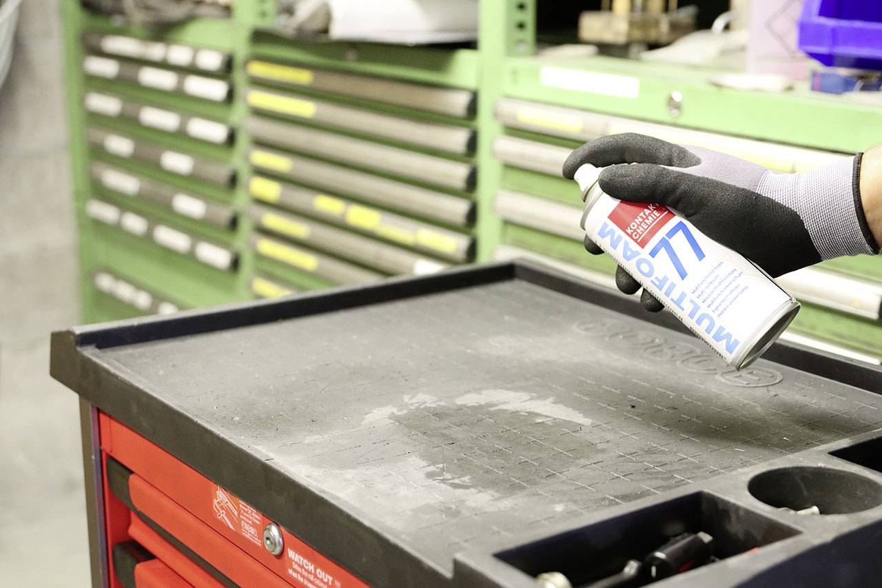 A gloved person is spraying a spray onto a tool trolley in a workshop with many drawers.