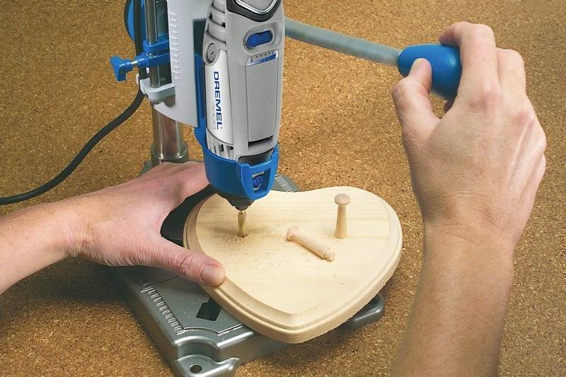 A person is using a pillar drill to work on a heart-shaped wooden board. A wooden dowel is resting on the board.