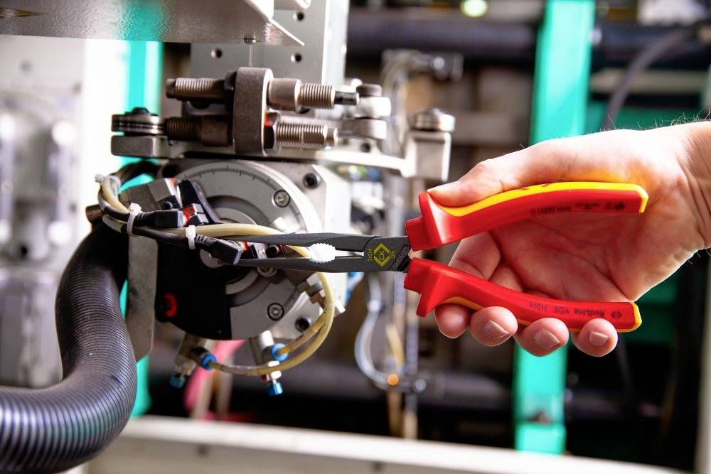 A hand holds a red pair of pliers working on a technical device. Machines and cables are visible in the background.