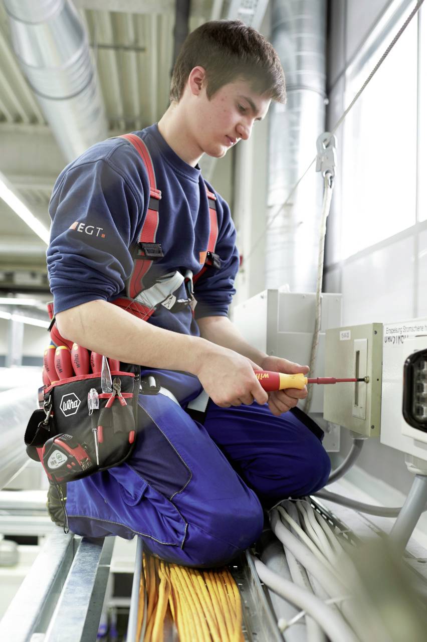 A young man in workwear is sitting on a cable trunking and working on a wall with a screwdriver and electrical equipment.