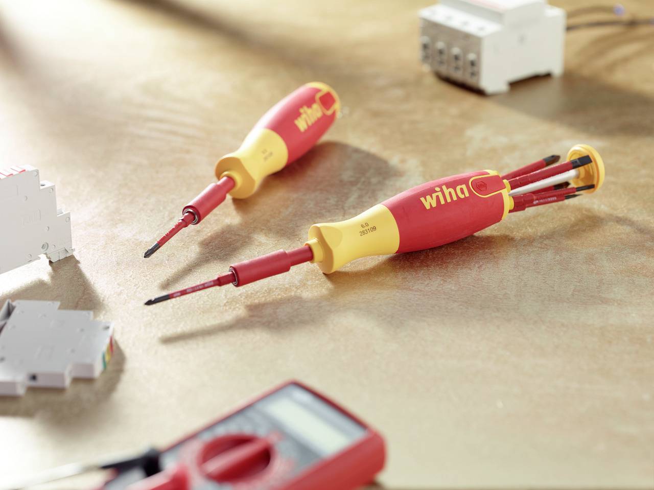 Two red screwdrivers with yellow handles are lying on a table, next to an electrical measuring device. Technical accessories in the background.