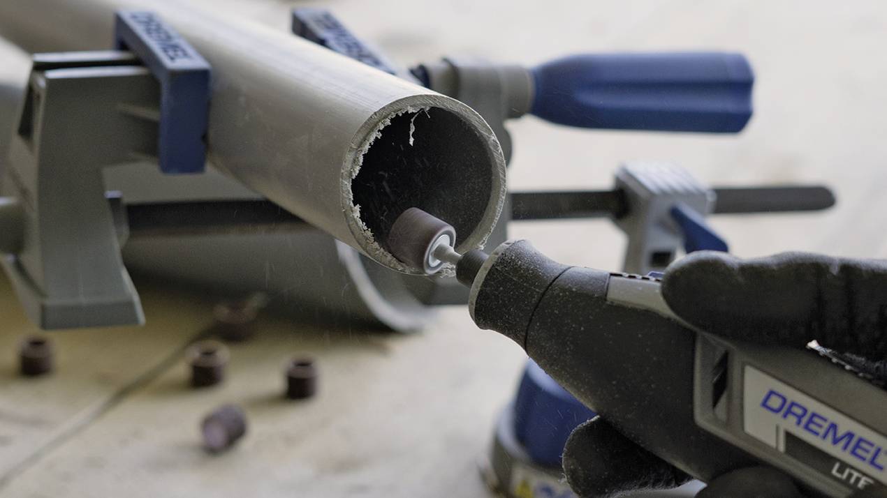 'Power tool cutting plastic pipe, held by G-clamps. Grinding attachment finishing edge of the pipe, shavings visible.'
