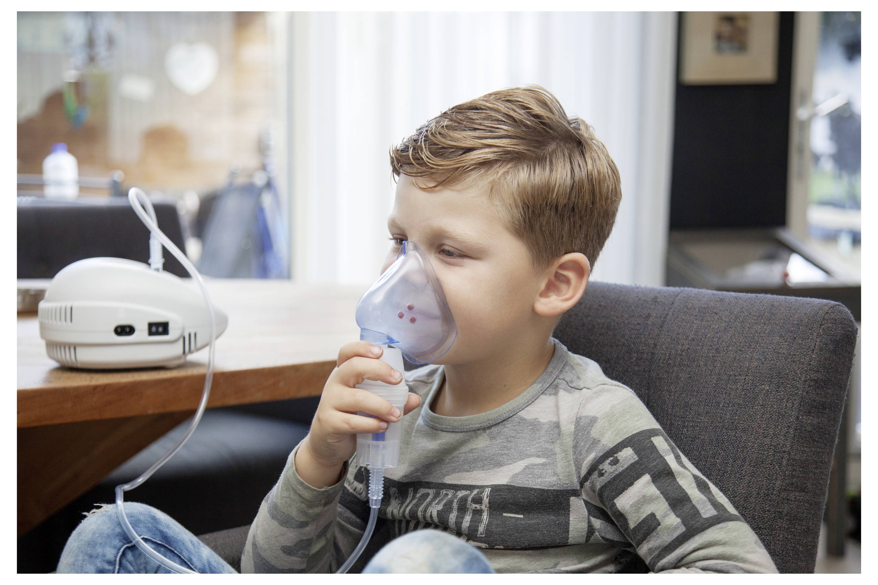 A young boy sits on a chair using a nebulizer with a mask over his face, indoors. The nebulizer is connected to a machine on a table.