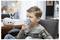 A young boy sits on a chair using a nebulizer with a mask over his face, indoors. The nebulizer is connected to a machine on a table.