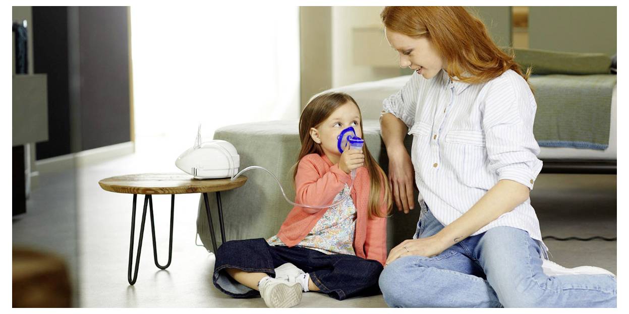 A mother watches her young child using a nebulizer at home, sitting on the floor together. The child inhales medicine from the device.